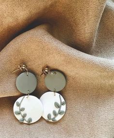 two circular earrings with leaves on them sitting on a fabric covered tablecloth, one is white and the other is grey