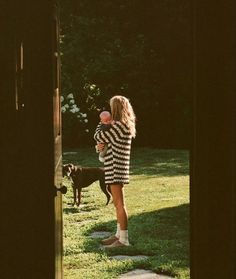 a woman holding a baby in her arms while standing next to a dog on the grass