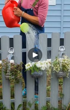 a woman watering flowers in front of a fence with water pouring from a red watering can