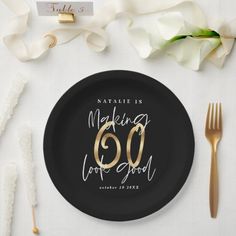 a black and gold plate with the number 60 on it next to white flowers, forks and napkins