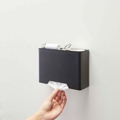 a hand holding a tissue dispenser over a white wall with a black box on it