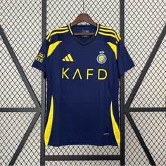 a blue and yellow soccer jersey hanging on a wall next to a metal fence with the word kafd printed on it