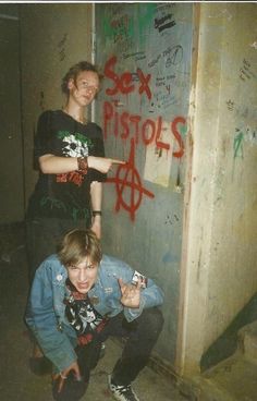 two people standing next to each other with graffiti on the wall behind them and one pointing at something