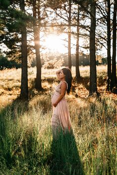 a pregnant woman standing in tall grass surrounded by pine trees at sunset with the sun shining behind her