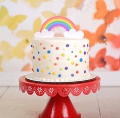 a white cake with rainbow decoration on top sitting on a red stand in front of a colorful wall