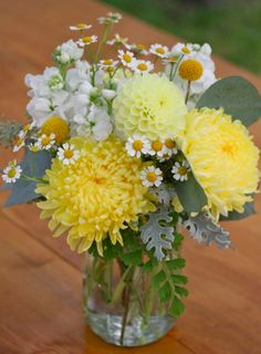 a glass vase filled with yellow and white flowers on top of a wooden table next to grass