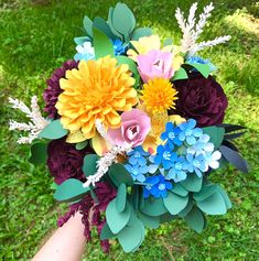 a bridal bouquet made with paper flowers and leaves in someone's hand on the grass