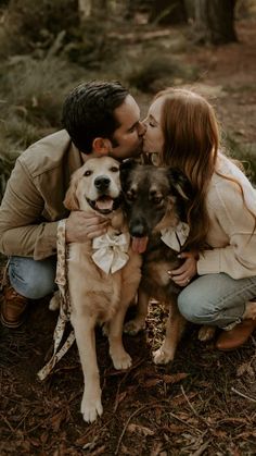 a man and woman kissing while holding two dogs in the woods with their faces close to each other