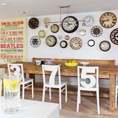 there are many clocks on the wall above the table in this dining room with white chairs