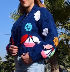 a woman standing in front of a palm tree wearing a blue sweater with boats and clouds on it