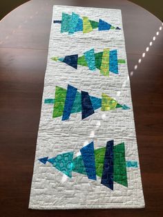 a quilted table runner with blue, green and yellow triangles on the top is sitting on a wooden table