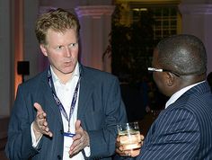 two men in suits talking to each other while holding drinks and looking at the camera