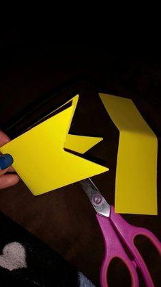 someone cutting out yellow paper with scissors on top of a black surface next to a pair of pink scissors