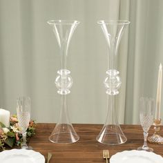 two clear vases sitting on top of a wooden table next to plates and candles