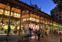 many people are walking around in front of a building at night with the lights on