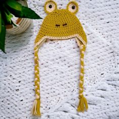 a crocheted frog hat on top of a white blanket next to a potted plant