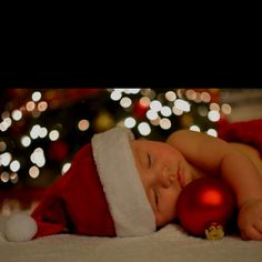 a baby wearing a santa hat sleeping next to a christmas tree with lights in the background