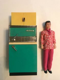 a doll standing next to a green and yellow refrigerator