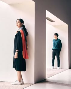 two people standing next to each other in front of a white wall and one is wearing a black graduation gown