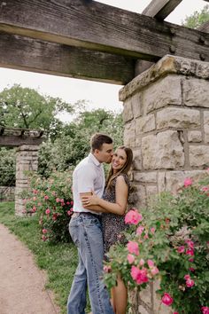 a man and woman standing next to each other near flowers in front of a stone wall