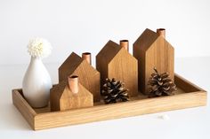 a wooden tray with pine cones and small houses on it, sitting next to a white vase