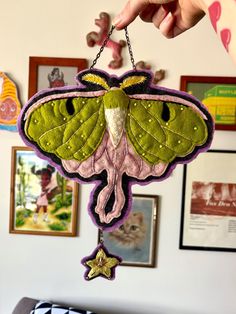 a hand holding a green and purple moth hanging from a chain with pictures on the wall behind it