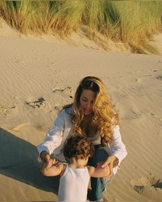 a woman sitting on top of a sandy beach next to a little boy holding his hand