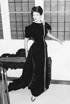 an old photo of a woman standing in front of a table wearing a long black dress