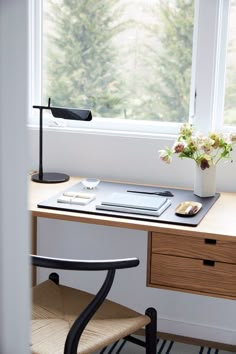 a desk with a laptop and flowers in a vase on it next to a window