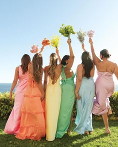 four women in dresses are holding flowers and looking at the ocean