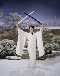 a woman holding two swords in her hands while standing on snow covered ground next to bushes