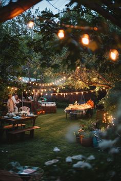 an outdoor party with lights strung from the trees and people sitting at tables in the grass