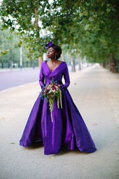 a woman in a long purple dress standing on the side of a tree lined street