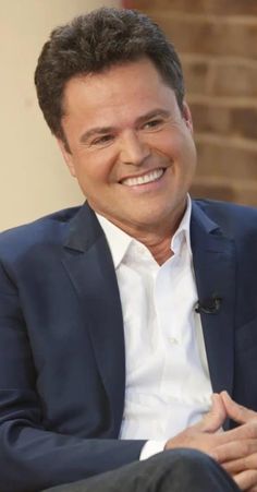 a smiling man in a blue suit and white shirt sitting on a chair with his hands crossed