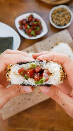 two hands holding up a piece of sushi with rice and tomatoes on it, next to other plates of food