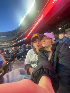 two women sitting in the stands at a baseball game, one is holding her arm around the other's shoulder