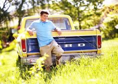 a man leaning on the back of a pickup truck in a grassy field with trees