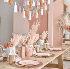 a dining room table set with pink and white plates, napkins, vases and tassels hanging from the ceiling