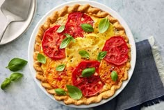 a pie with tomatoes and basil on top sits on a table next to a spoon