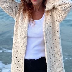 a woman standing on the beach with her arms behind her head, wearing a white sweater and black pants