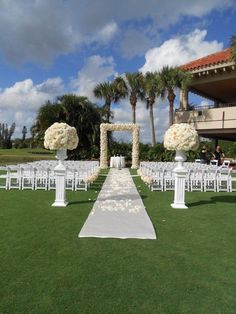 an outdoor ceremony setup with white chairs and flower arrangements on the aisle, in front of palm trees