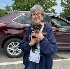 an older woman holding a small dog in her arms while standing next to a parked car