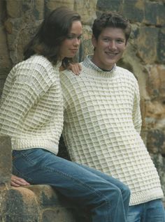 a man and woman sitting next to each other in front of a stone wall wearing sweaters