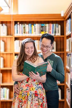 a man and woman standing next to each other in front of bookshelves holding an open book