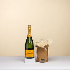 a bottle of champagne next to a brown paper bag on a white table with a tan wall in the background