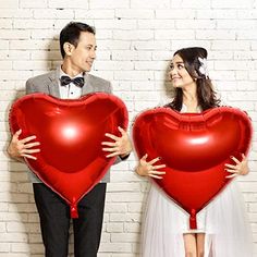 two people holding red heart shaped balloons in front of a brick wall