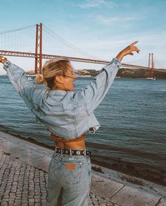 a woman standing next to the water with her arms in the air while wearing jeans