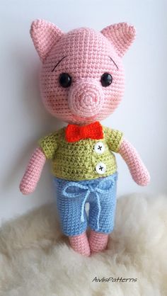 a crocheted pig with a yellow shirt and blue pants is standing on a fluffy white surface