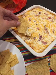 a hand dipping a tortilla chip into a casserole with cheese and bacon