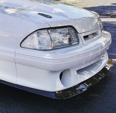 the front end of a white car parked in a parking lot next to a tree